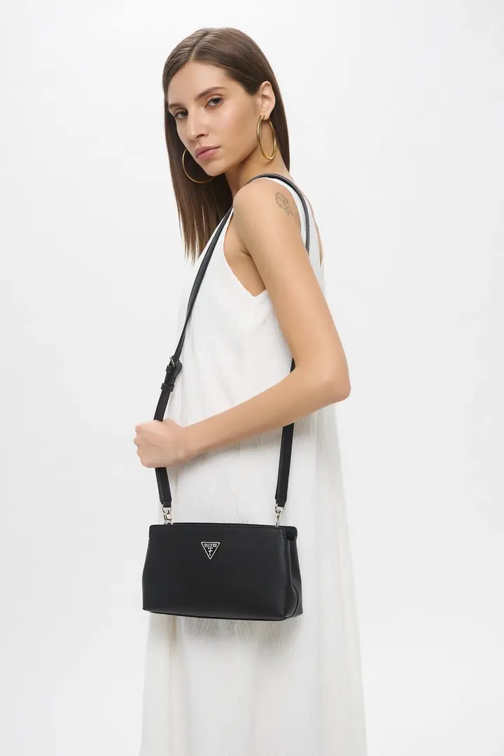 A model in a white dress wears a black leather crossbody bag and turns to the camera.